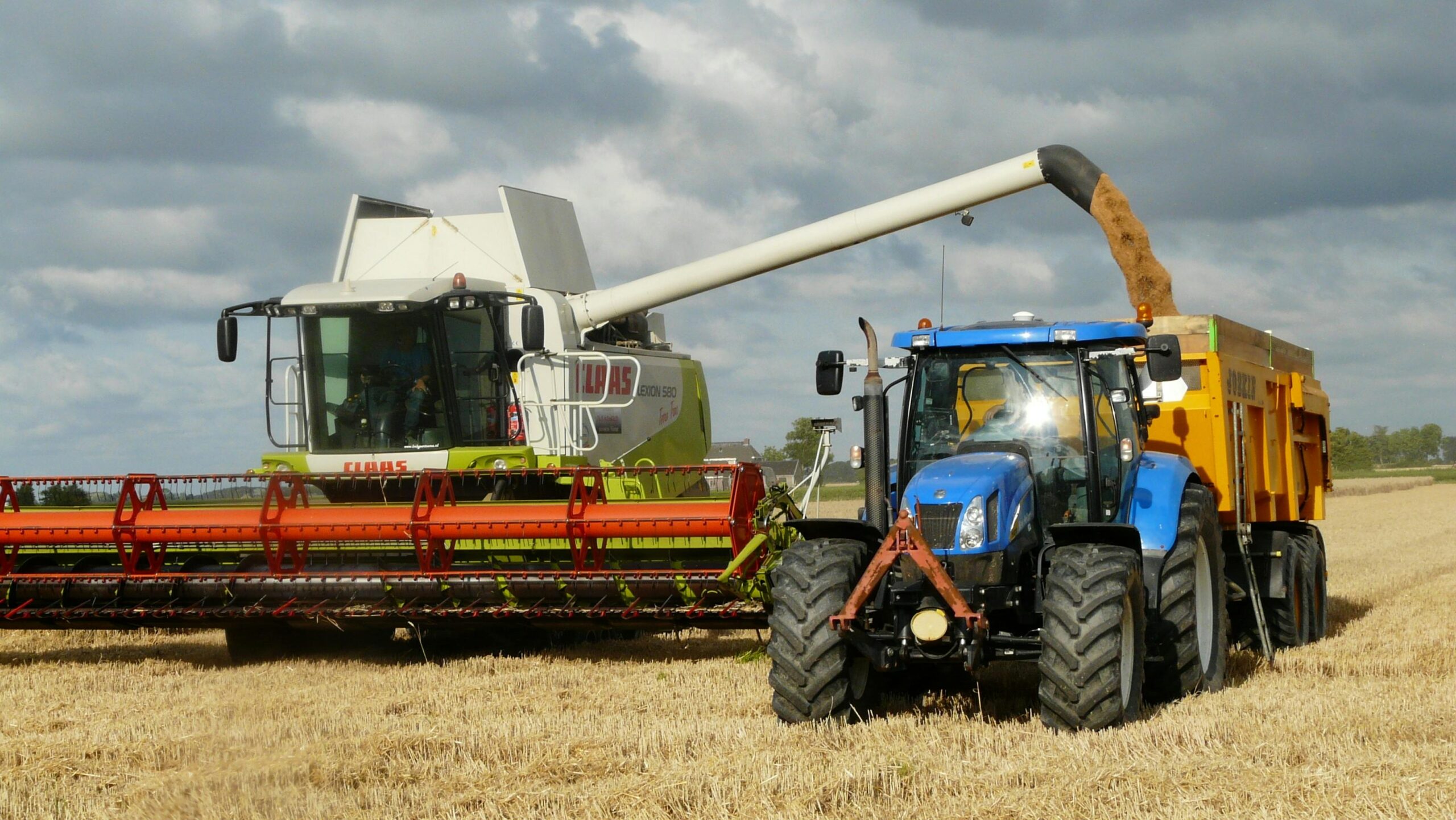 A modern combine harvester and tractor working together to harvest wheat in a sunny outdoor field.